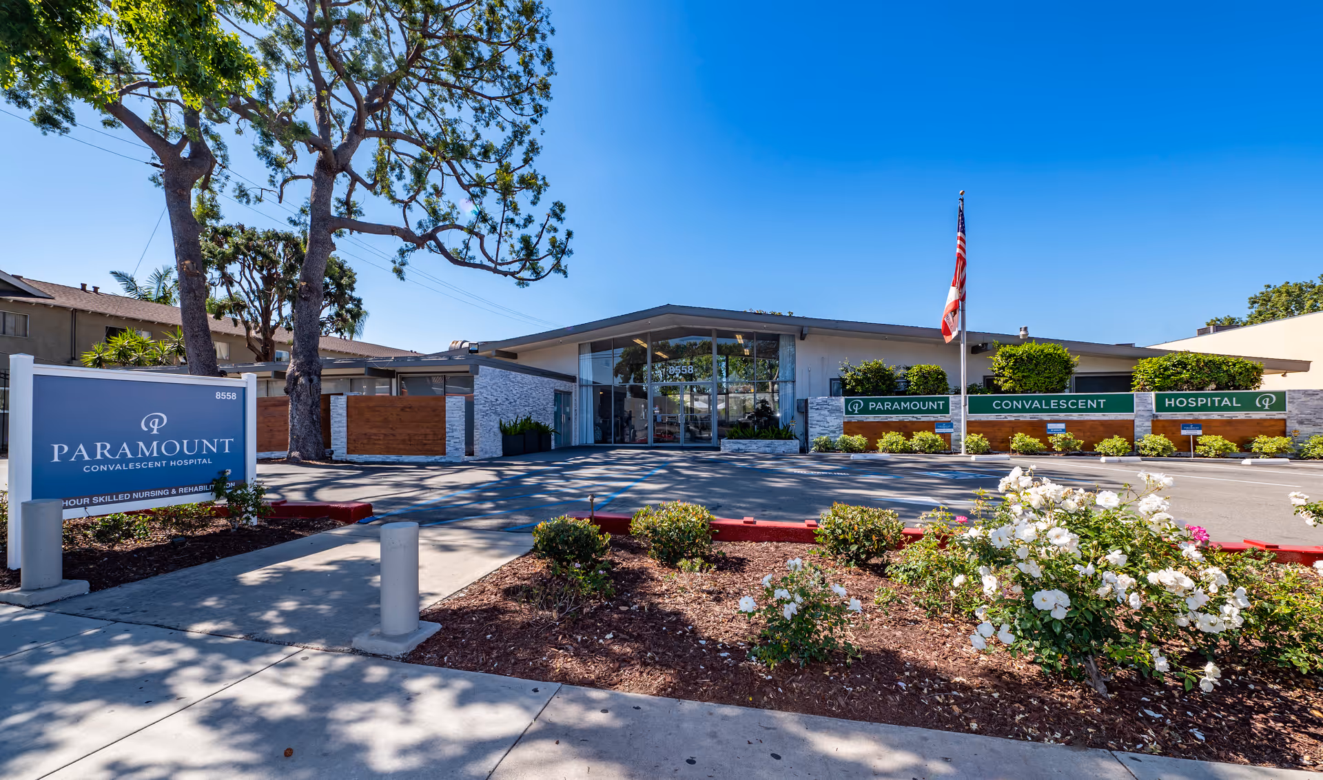 Front exterior view of Paramount Convalescent Hospital building with a clear blue sky, landscaped bushes and flowers, an American flag on a flagpole, and a large sign displaying the facility's name near the sidewalk.