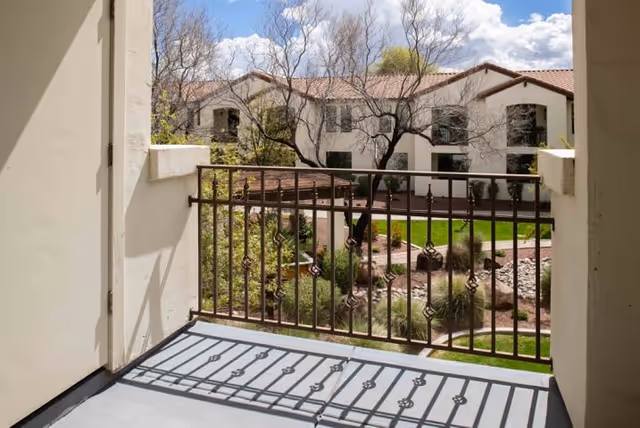 View from a balcony with a decorative metal railing overlooking a landscaped courtyard with grass, trees, and a walking path, surrounded by a two-story building with white walls and red-tiled roofs under a partly cloudy sky.