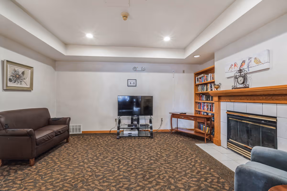A cozy living room with a brown patterned carpet, a brown leather couch on the left, a flat-screen TV on a glass stand in the center against the wall, a wooden bookshelf filled with books on the right, a wooden side table, and a fireplace with a wooden mantle decorated with a clock and a painting of birds above it.