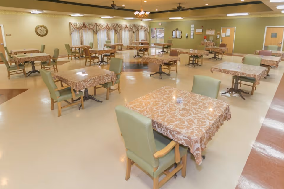 A spacious dining room with multiple square tables covered with patterned tablecloths. Each table is surrounded by green cushioned chairs. The room has a beige and green color scheme with a clock on the wall, ceiling fans, and windows with decorative curtains.