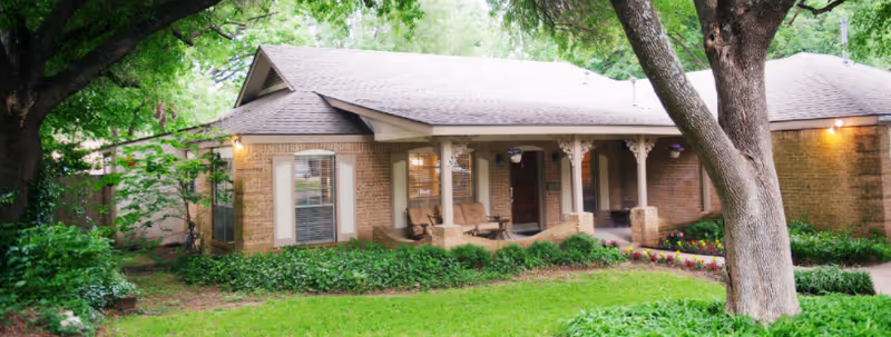 Front exterior view of a single-story brick house with a covered porch featuring seating, surrounded by green grass, plants, and large trees.