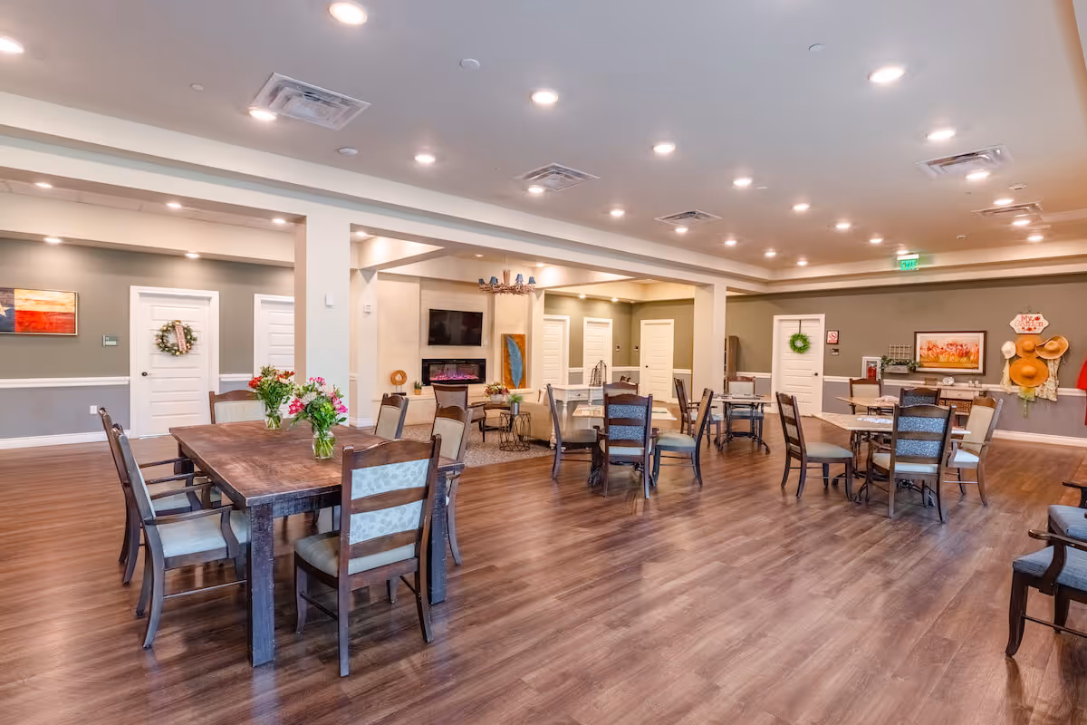 Spacious and well-lit common area with multiple wooden tables and chairs arranged on a polished wooden floor. The room features a modern electric fireplace with a TV mounted above it, decorative wall art, and floral arrangements on the tables. The walls are painted in neutral tones with white doors and trim, and the ceiling has recessed lighting.