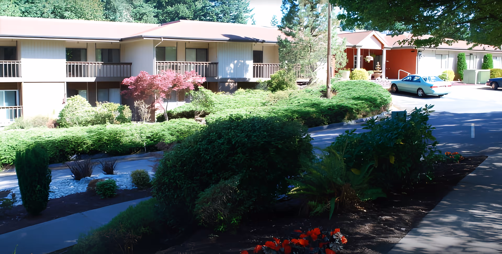 Exterior view of a senior living facility with a two-story building featuring balconies, surrounded by well-maintained landscaping including bushes, trees, and flowers. A driveway and parked cars are visible near the entrance.