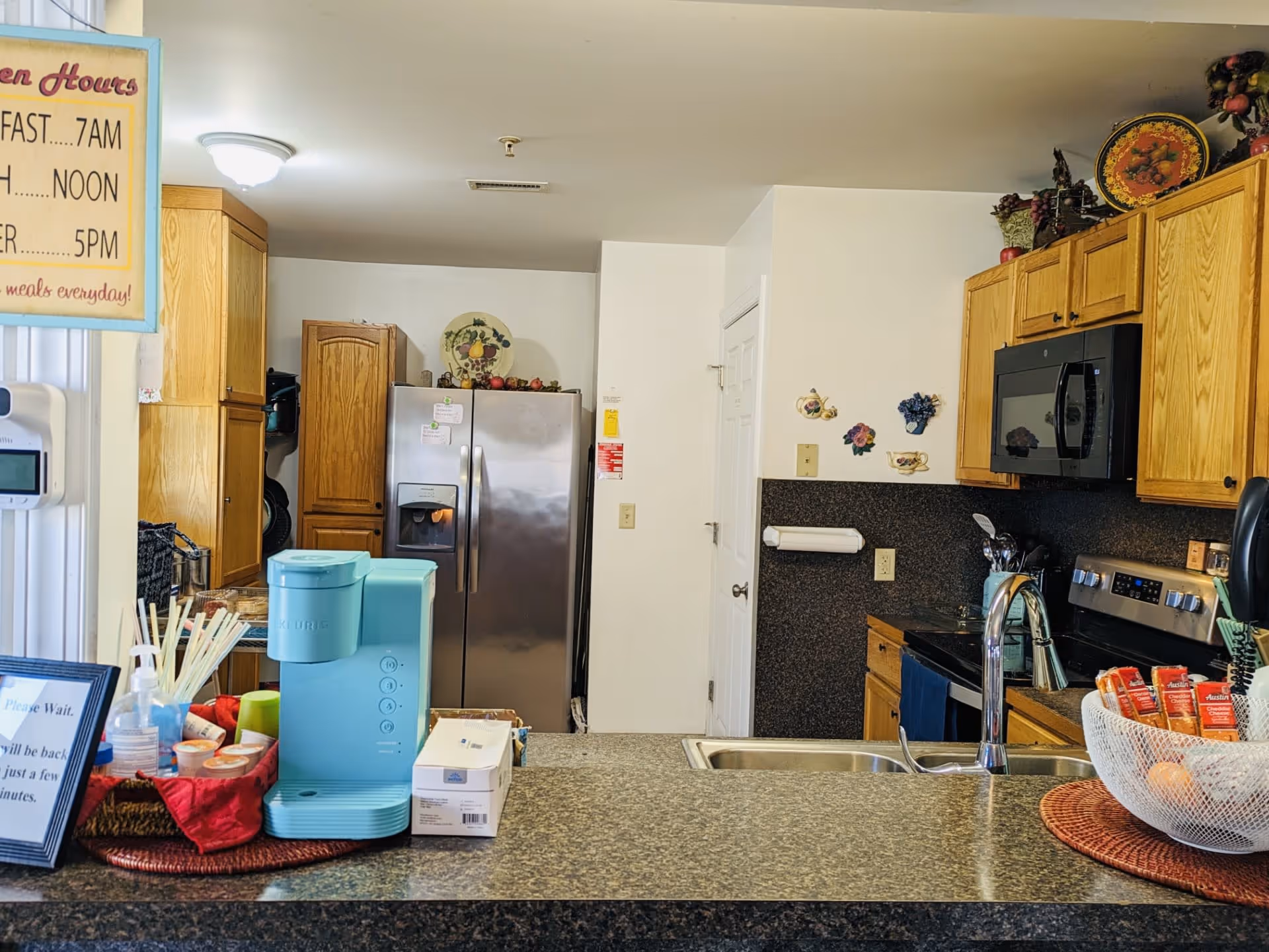 Interior view of a kitchen with wooden cabinets, a stainless steel refrigerator, a microwave, and a stove. A countertop with a sink and a blue coffee machine is in the foreground. Various kitchen items and decorations are visible, including a bowl with snacks and a sign displaying open hours.