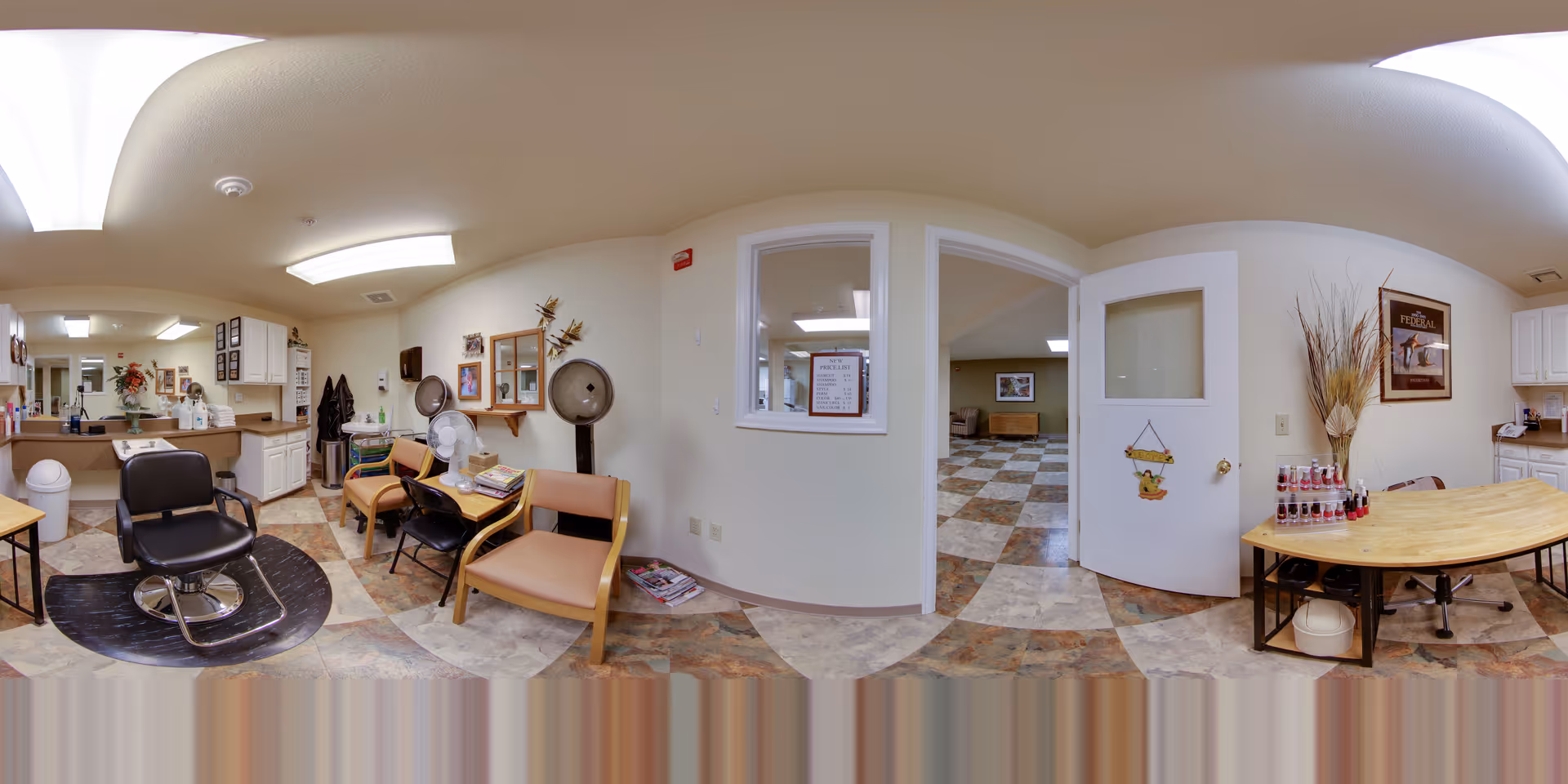 Interior view of a senior living facility's salon area with a black salon chair on a protective mat, beige chairs along the wall, hair drying equipment, and a small table with decorative items and beverages. The room has tiled flooring, white walls, and ceiling lights. A door and window open into an adjacent room with similar flooring and a framed picture on the wall.