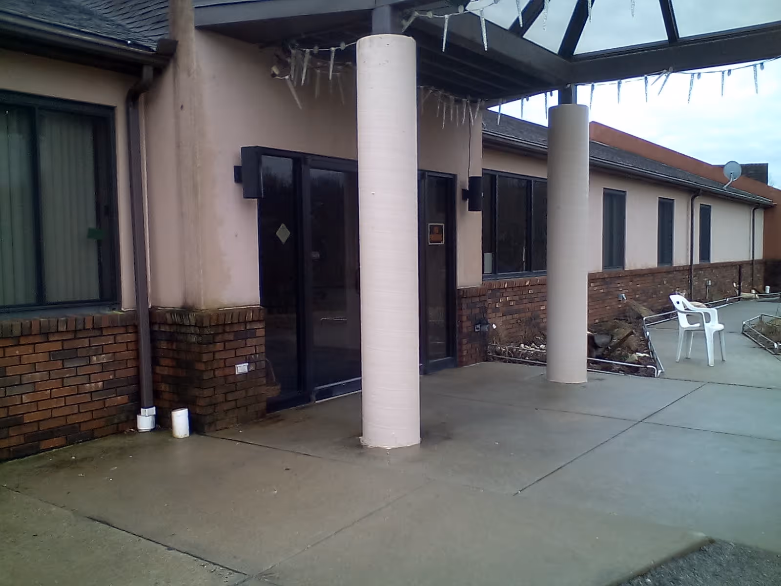 Exterior view of a building entrance with two large round columns supporting a glass canopy. The building has brick and beige walls with several windows. There are two white plastic chairs on the concrete patio near the entrance.