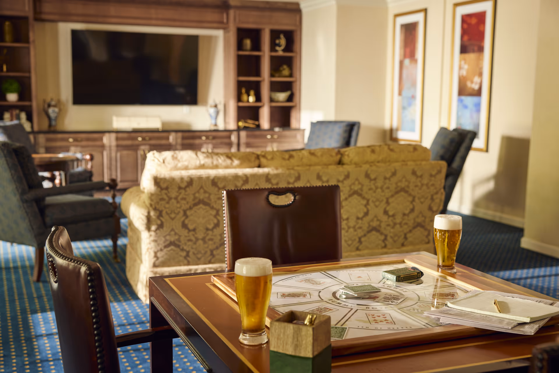 A cozy living room area with a patterned beige sofa, blue armchairs, and a large flat-screen TV mounted on a wooden entertainment center. In the foreground, there is a game table with playing cards, two glasses of beer, and a notepad with a pen. The room has blue carpet with a yellow dot pattern and framed artwork on the walls.