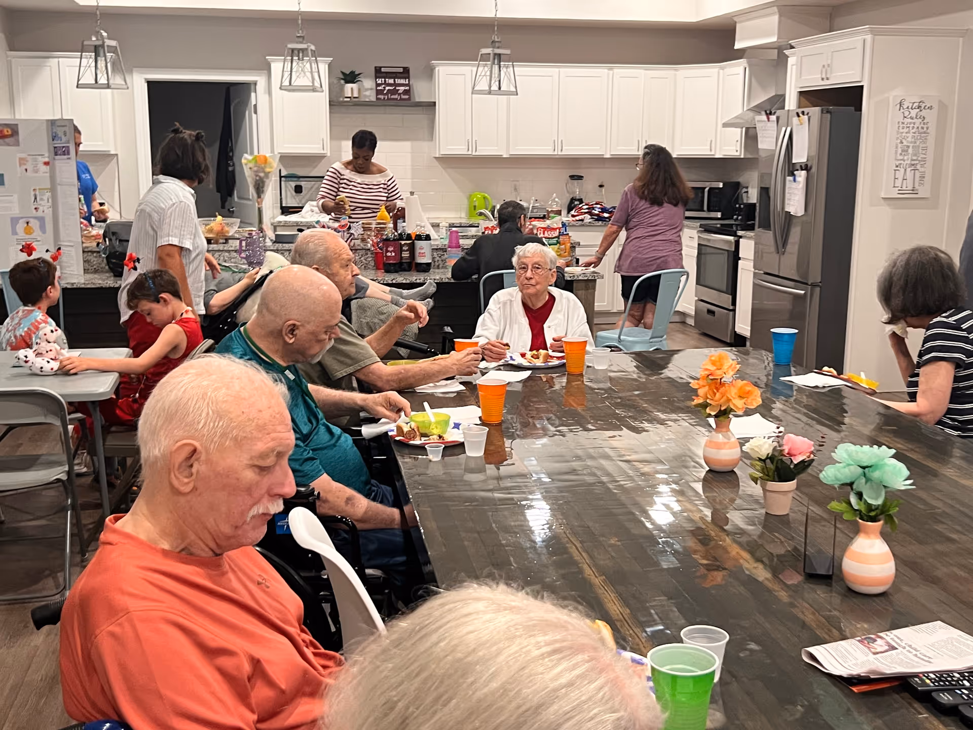 Residents and staff gathered around a large communal dining table in a kitchen/dining area, eating and socializing.