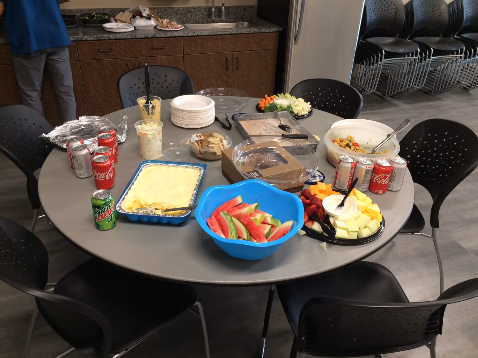 A round table in a room with various food items and drinks arranged on it, including watermelon slices in a blue bowl, a tray of yellow casserole, a vegetable platter with dip, a fruit platter, several cans of soda, and containers of salad and other dishes. There are black chairs around the table and a countertop with cabinets in the background. A person wearing a blue shirt and gray pants is partially visible standing near the counter.