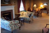 A cozy living room area with floral upholstered sofas, a small round wooden table, a fireplace with a white mantle, and a dining table with chairs in the background. The room is softly lit with lamps and natural light coming through a window with red curtains.