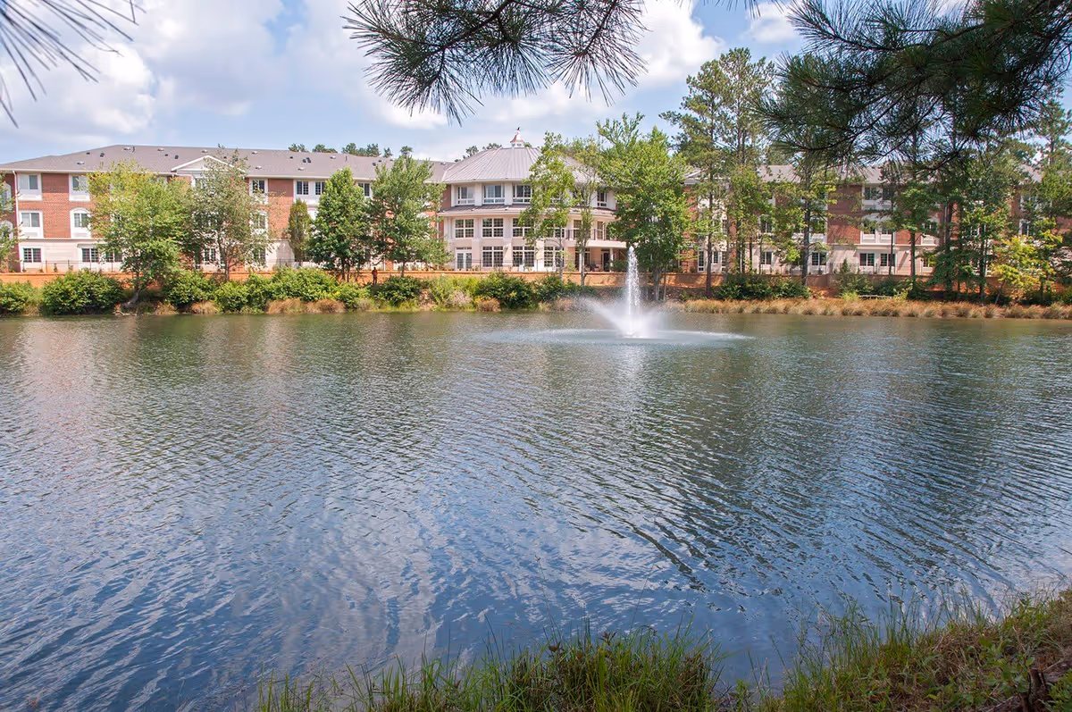 A large pond with a water fountain in the center, surrounded by grass and trees. In the background, there is a multi-story brick and white building partially obscured by trees under a partly cloudy sky.