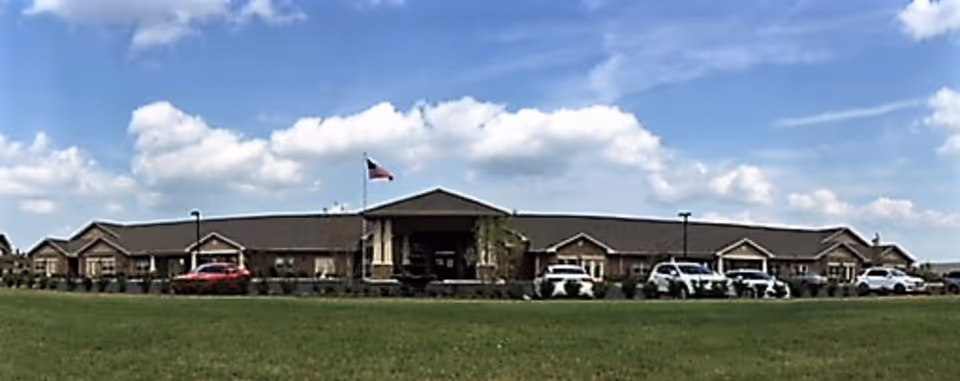 Wide exterior view of Culpepper Place, a single-story building with a central entrance featuring a covered porch and an American flag flying above. Several cars are parked in front of the building under a partly cloudy blue sky.