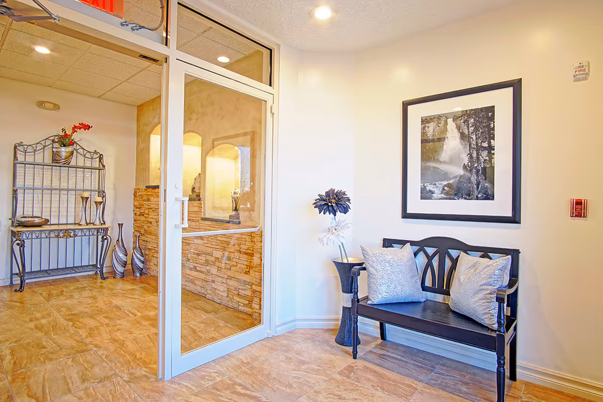 A bright and clean interior corner of a facility with a black wooden bench adorned with two silver pillows, a tall decorative vase with large artificial flowers, a framed black and white waterfall photograph on the wall, and a glass door leading to another room with stone wall accents and decorative metal furniture.