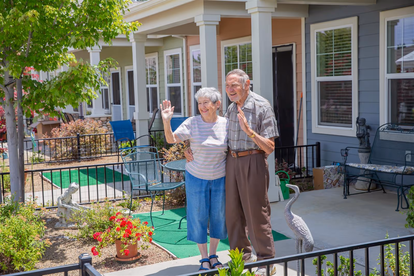 An elderly couple standing outside a residential building, smiling and waving. They are surrounded by garden plants, outdoor furniture, and decorative statues on a sunny day.