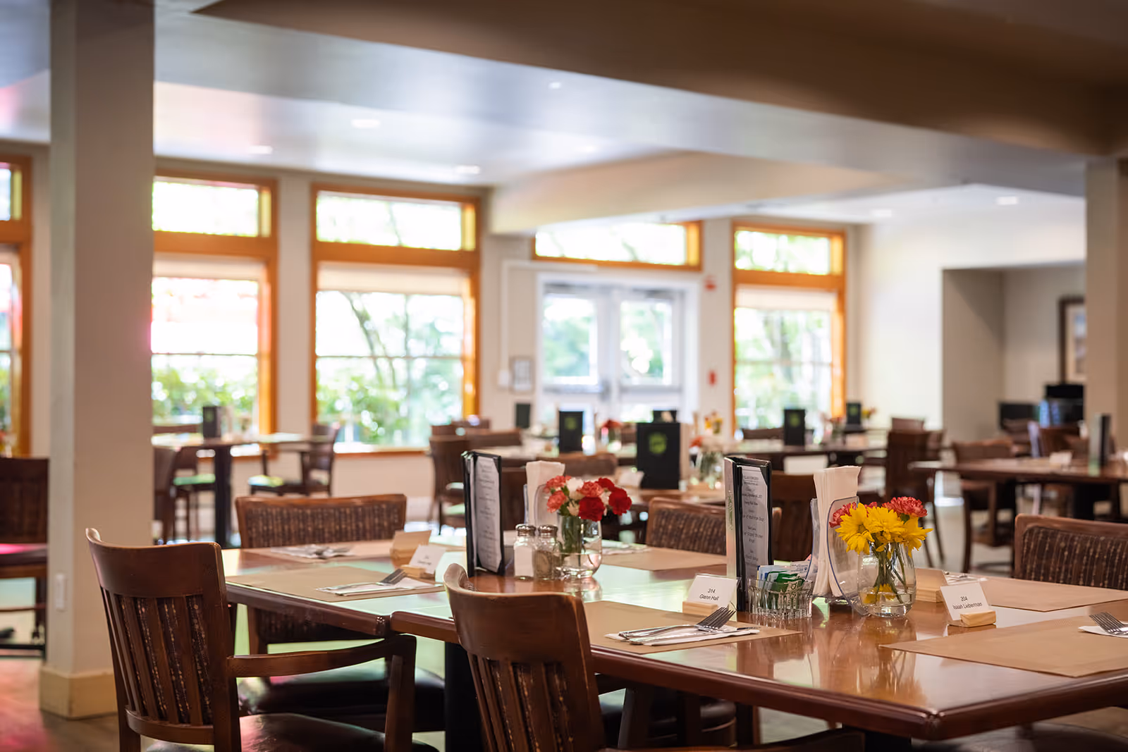 A bright and spacious dining room with wooden tables and chairs. Tables are set with placemats, menus, utensils, napkins, and small vases of colorful flowers. Large windows with wooden frames allow natural light to fill the room, creating a warm and inviting atmosphere.