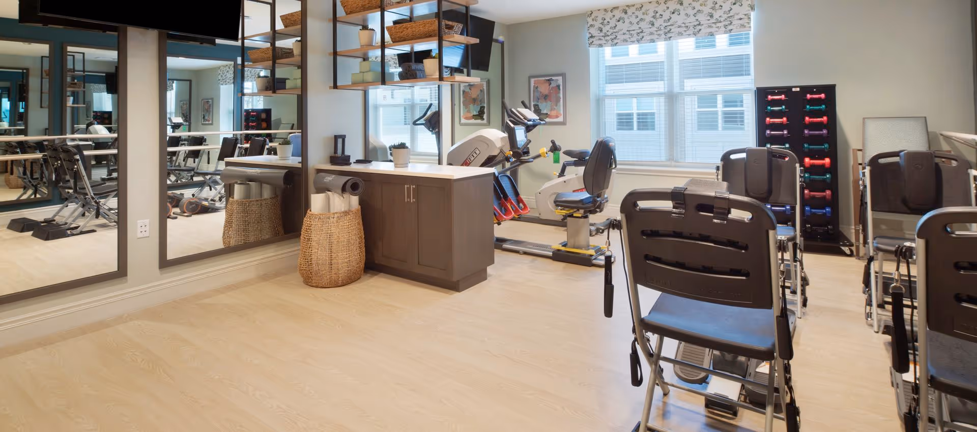 A bright fitness room with exercise bikes, several padded chairs, a dumbbell rack, large mirrors and storage cabinets.