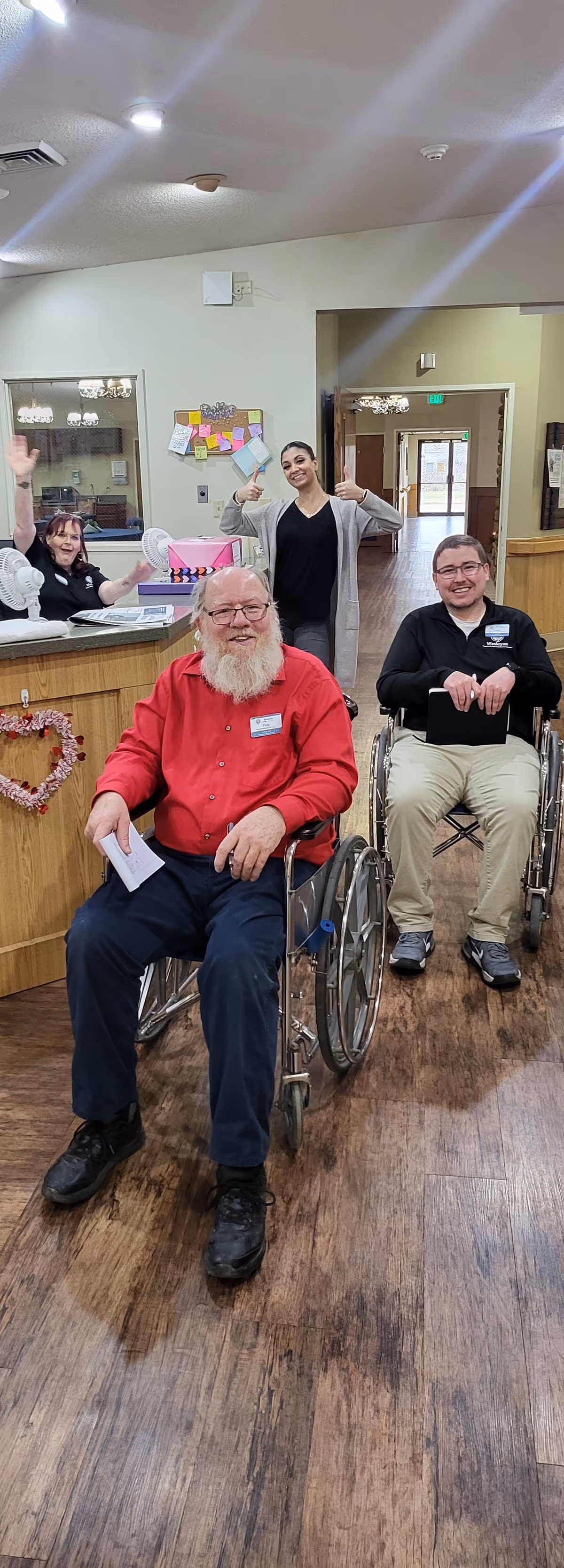 Two men in wheelchairs smiling in the foreground inside a senior living facility. Behind them, a woman stands giving two thumbs up, and another woman is seated at a reception desk waving. The setting appears to be a welcoming common area with wooden floors and light-colored walls.