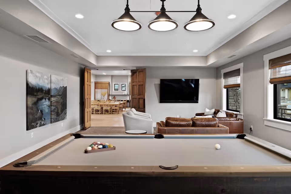 A modern recreational room featuring a pool table with balls arranged for a game, a seating area with brown leather sofas and a white armchair, a wall-mounted flat-screen TV, and large windows with wooden blinds. In the background, there is a dining area with a wooden table and chairs, visible through open double wooden doors. The room has neutral-colored walls and ceiling lights.