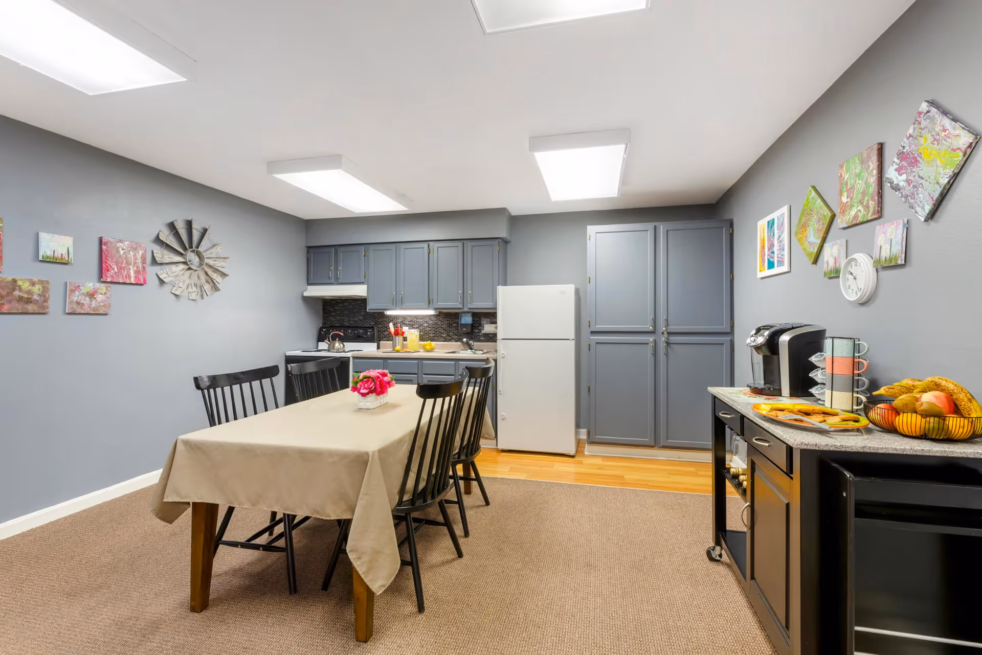 A kitchen and dining area with gray cabinets, a white refrigerator, and a table covered with a beige tablecloth surrounded by black chairs. The walls are decorated with colorful artwork and a clock. A countertop on the right holds a coffee maker, mugs, and a basket of fruit.