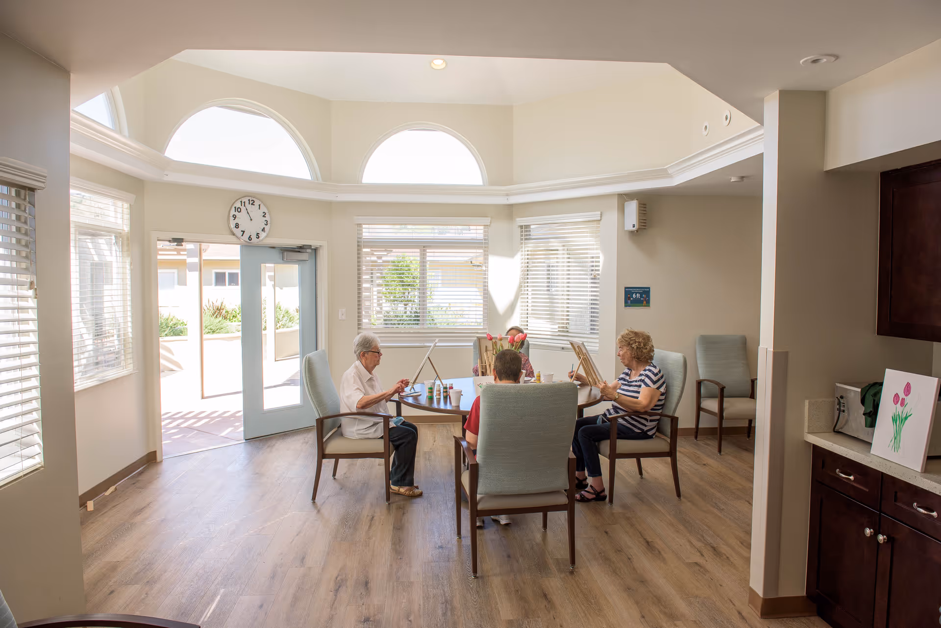A bright room with large windows and arched transom windows above a door. Four elderly people are seated around a round table engaged in painting activities with easels and paint cups. The room has wooden flooring and light-colored walls, with a clock above the door showing 10:10. There are additional chairs and a countertop with a painting of tulips on it.