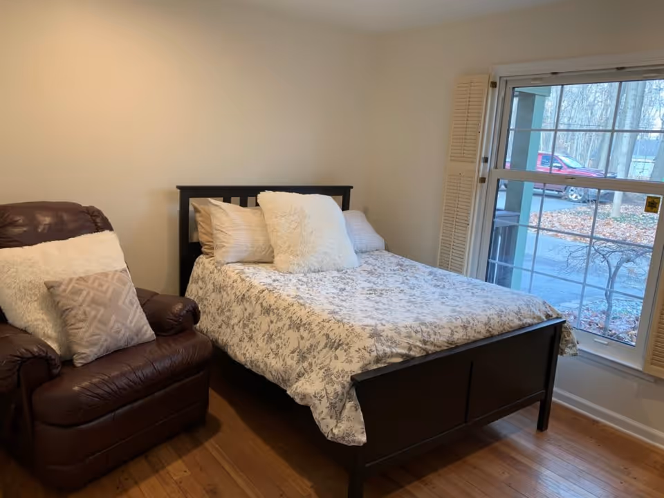 A cozy bedroom with a bed dressed in patterned bedding, a fluffy pillow, and a leather recliner next to a large window.