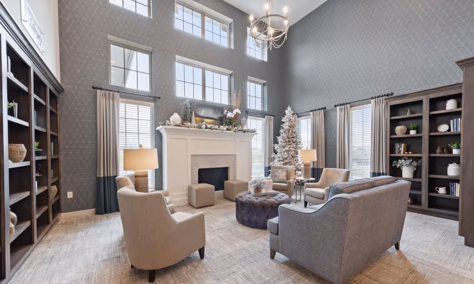 Bright, high-ceilinged communal living room with a fireplace, arranged seating, bookshelves and a decorated Christmas tree.
