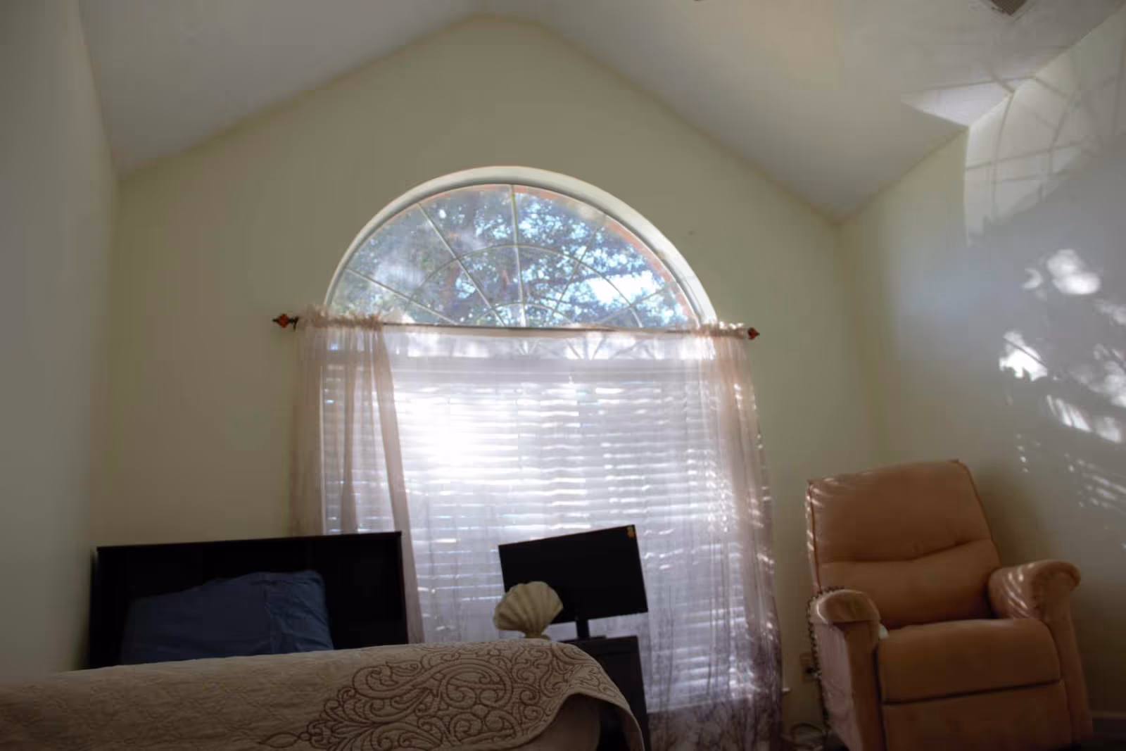 A bedroom with a bed covered in a patterned quilt, a blue pillow, a small table with a decorative shell, a monitor, and a beige recliner chair. The room has a large arched window with sheer curtains allowing sunlight to filter through.