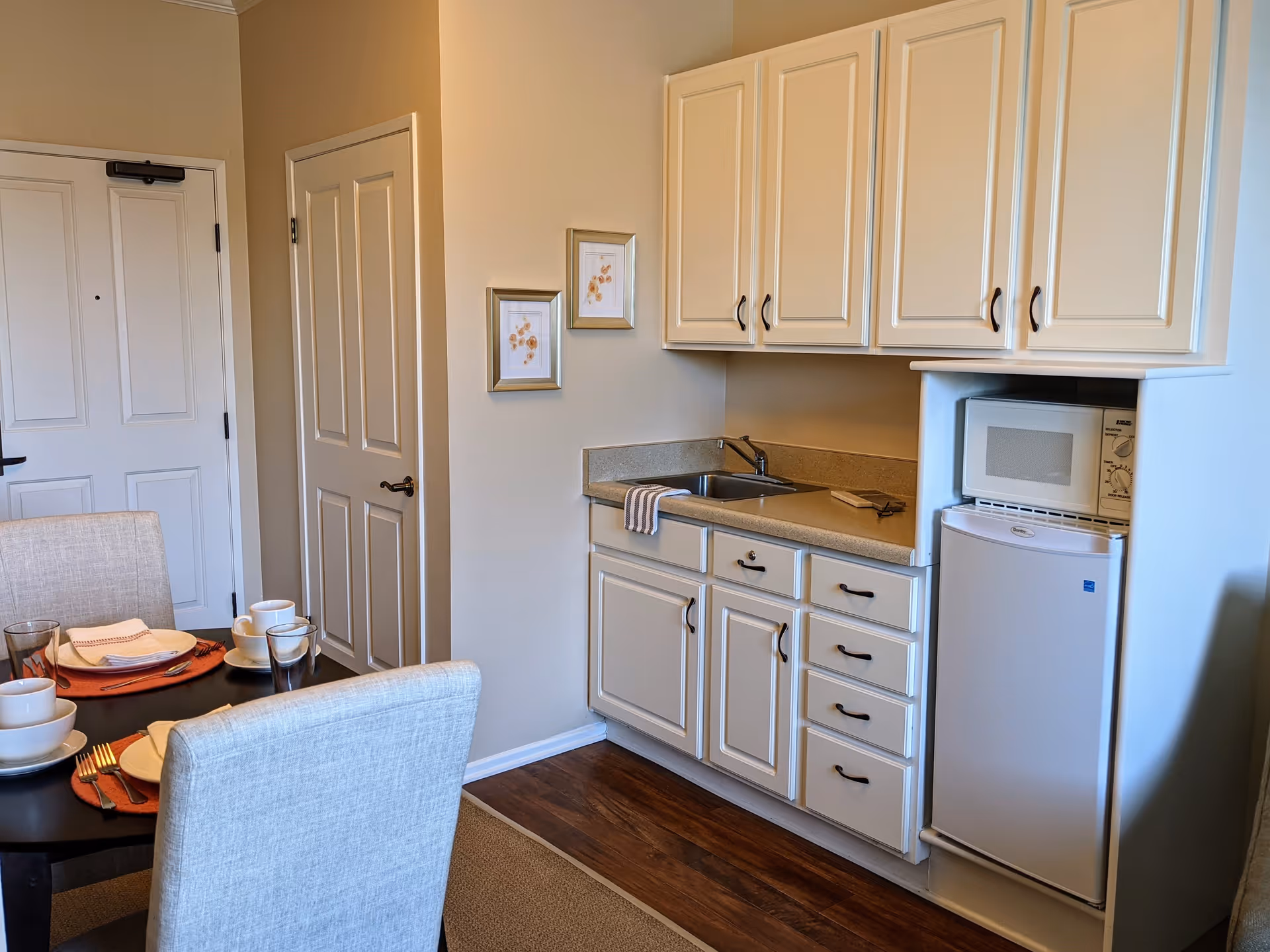 A small kitchen area with cream-colored cabinets, a countertop with a sink, a microwave, and a mini refrigerator. To the left, there is a dining table set with plates, cups, and utensils, and two upholstered chairs. The walls are light-colored with two framed pictures hanging. A door is visible in the background.