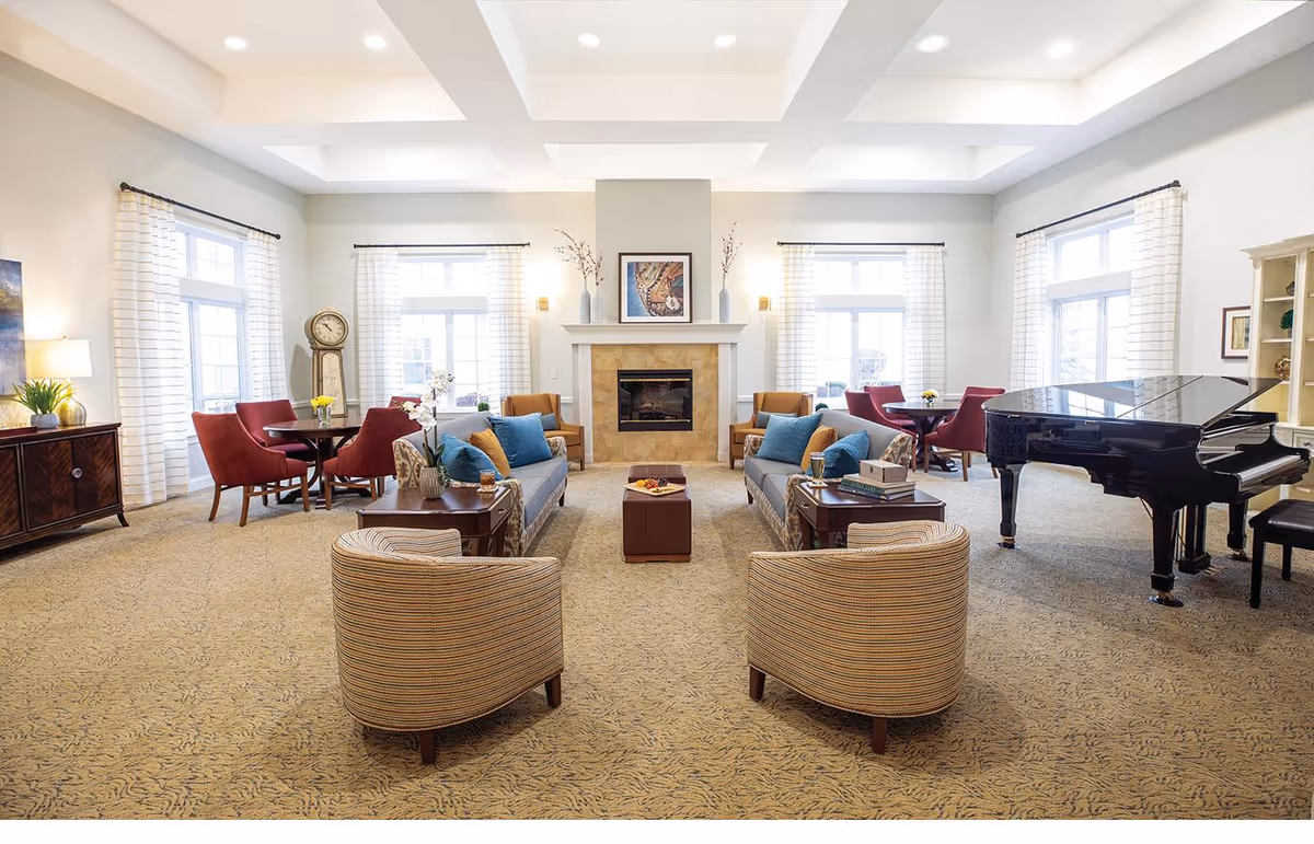 A spacious and well-lit living room with two blue sofas, two striped armchairs, a grand piano, and several small round tables with red chairs. The room features a central fireplace with artwork above it, large windows with white curtains, and a beige patterned carpet.