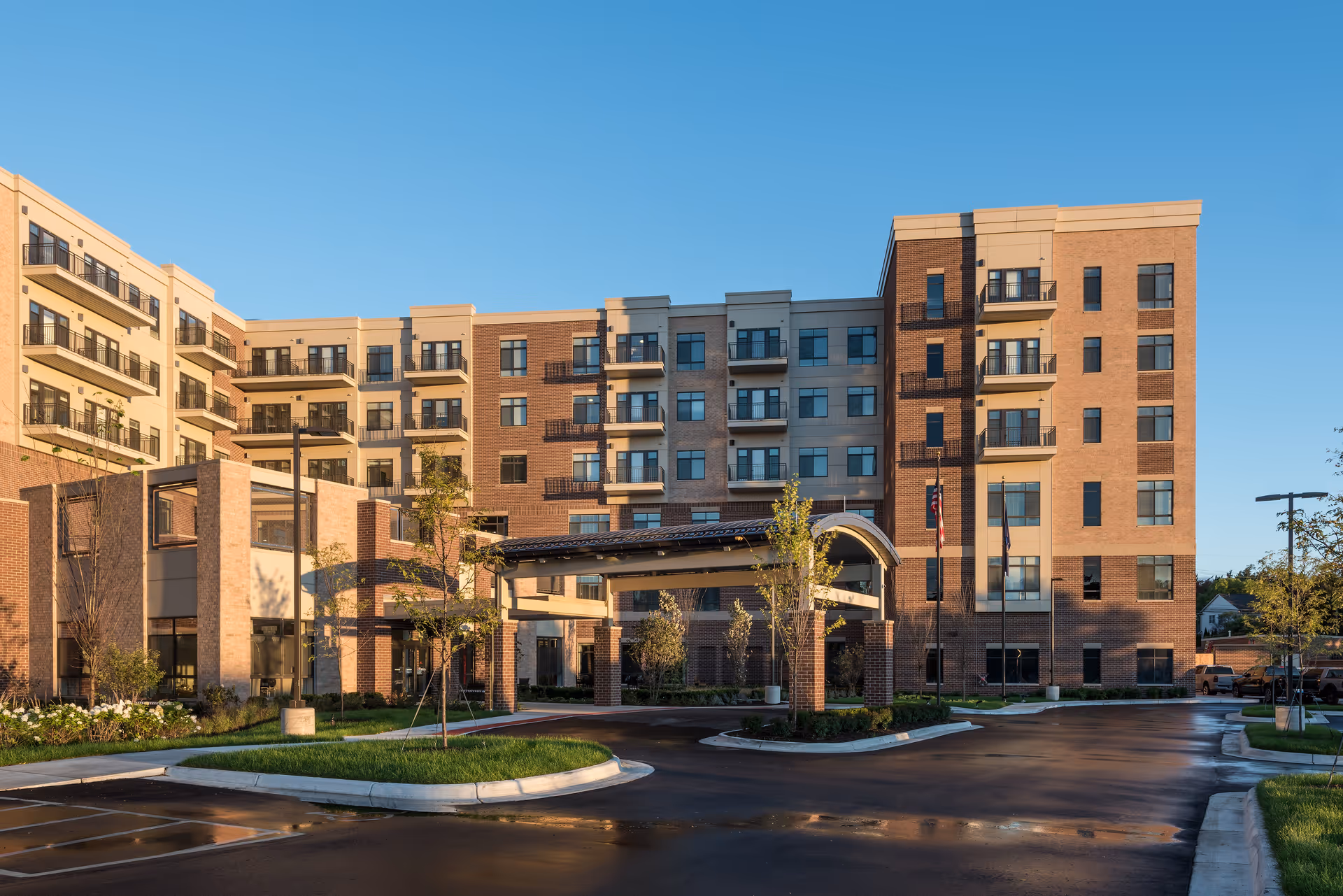Exterior view of a multi-story senior living facility building with balconies, a covered entrance, landscaped greenery, and a clear blue sky.