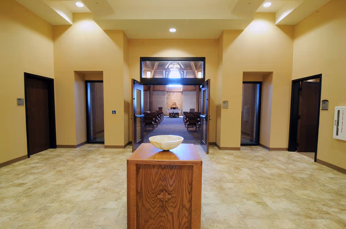 Interior view of a hallway leading to a chapel or worship room with wooden pews and an altar. The hallway has beige walls, tiled flooring, and a wooden stand with a bowl in the center. There are several doorways on either side of the hallway.