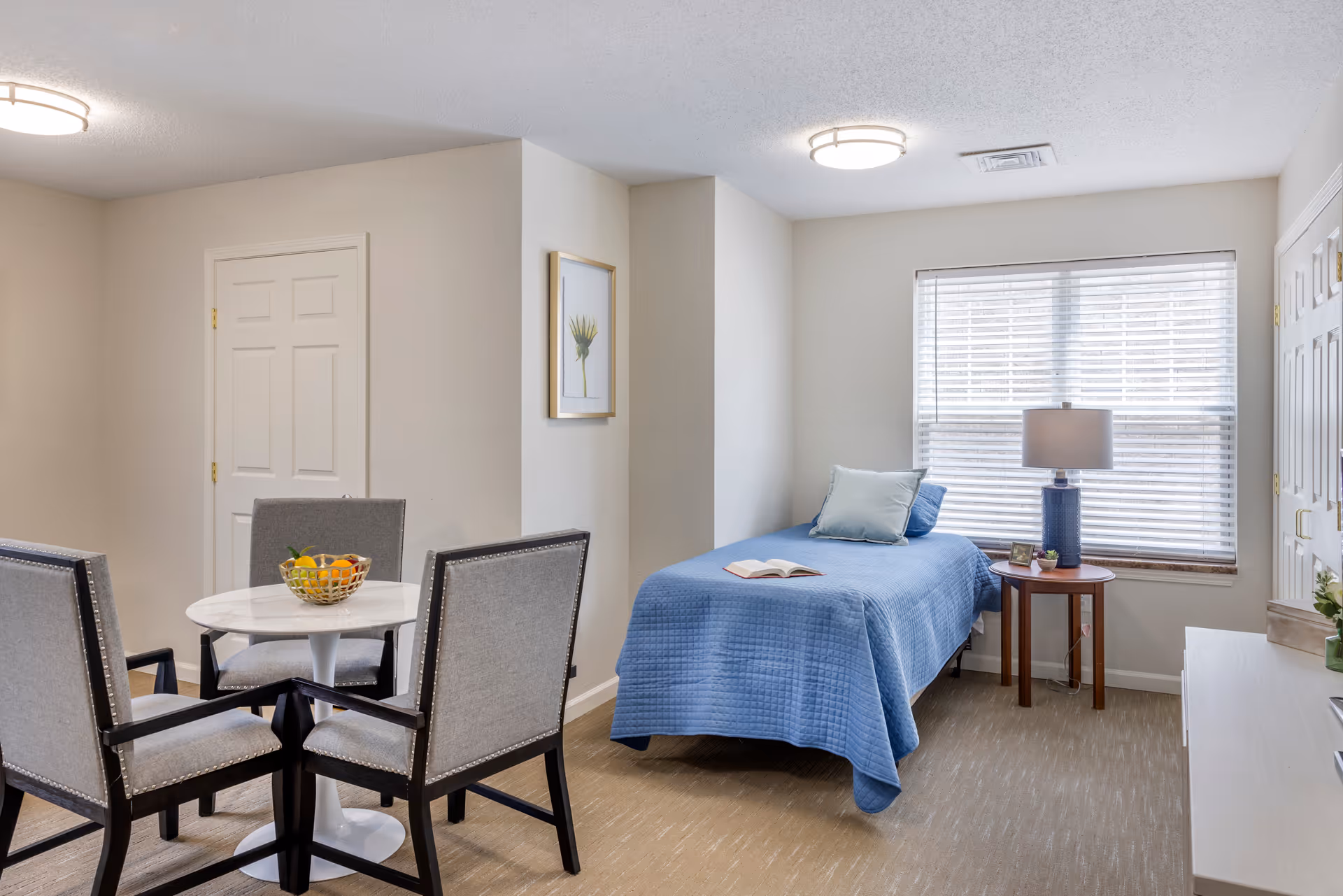 Light-filled studio bedroom with a single bed covered in a blue quilt, a small round dining table with four chairs, and a window with blinds.