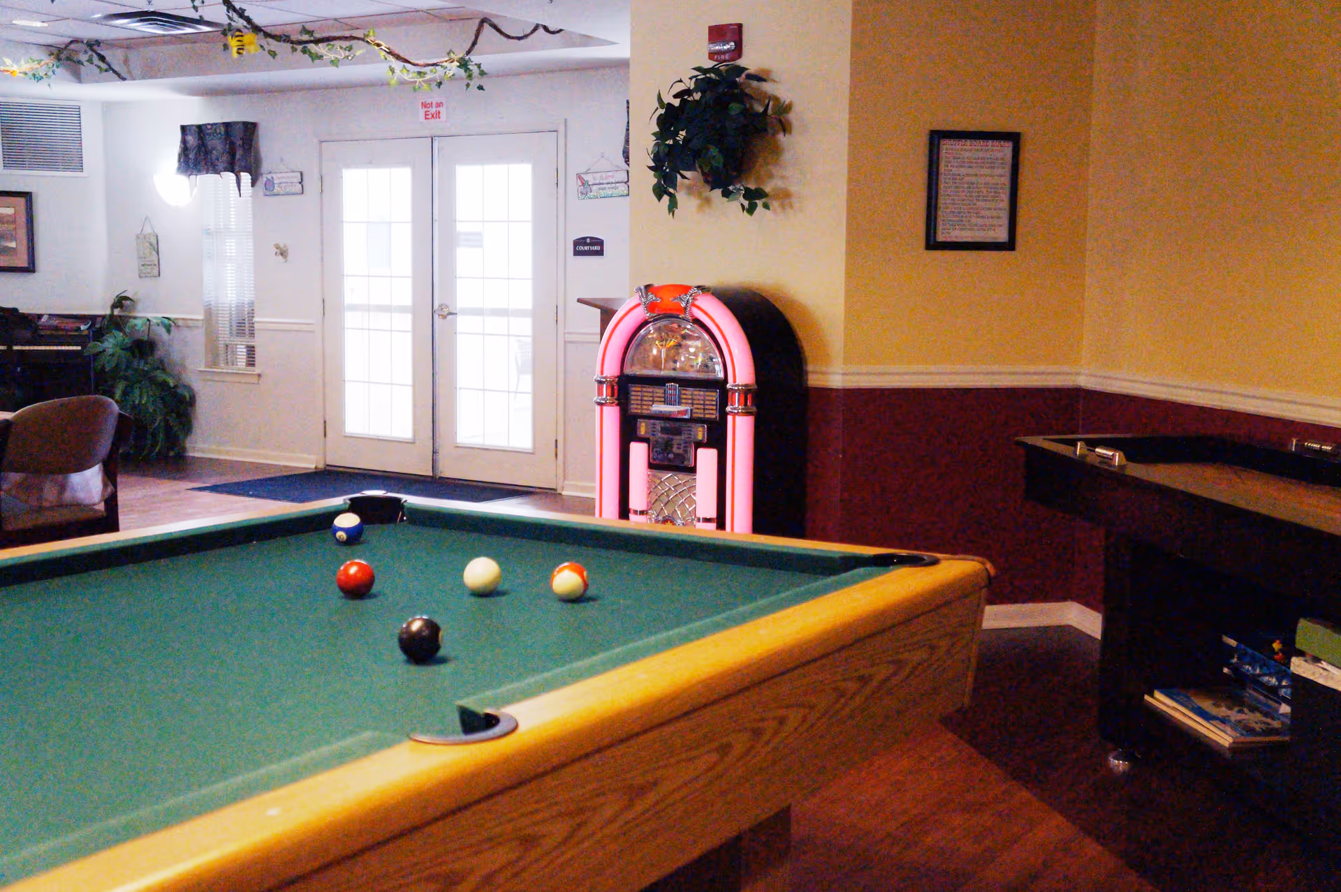 Interior view of a recreational room featuring a pool table with balls arranged on it, a vintage-style jukebox against the wall, a shuffleboard table, and seating area with chairs. The room has a warm color scheme with yellow and maroon walls, and double glass doors leading outside.