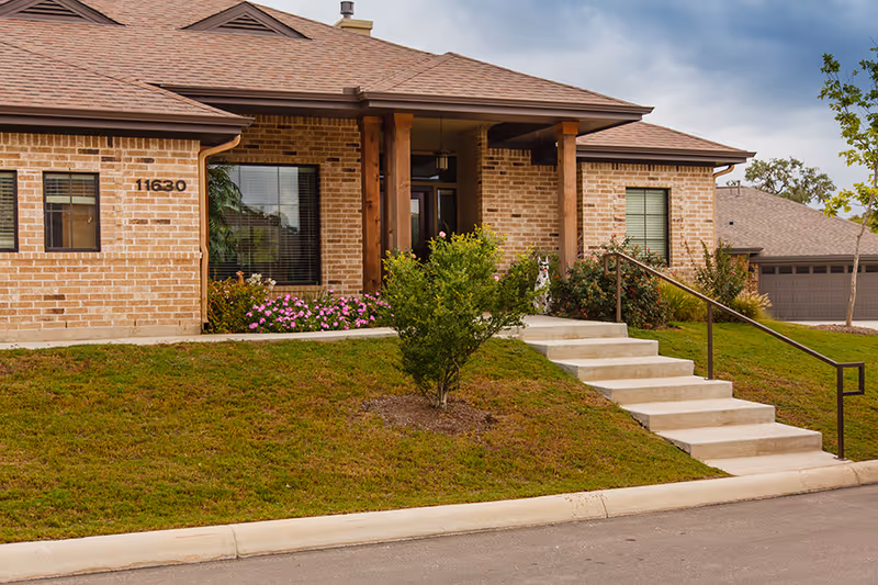 Exterior view of a single-story brick building with a sloped roof, a set of concrete stairs with a metal handrail leading up to a covered entrance supported by wooden pillars. There are windows with blinds, green grass, shrubs, and pink flowers in front of the building. The building number 11630 is visible on the wall.