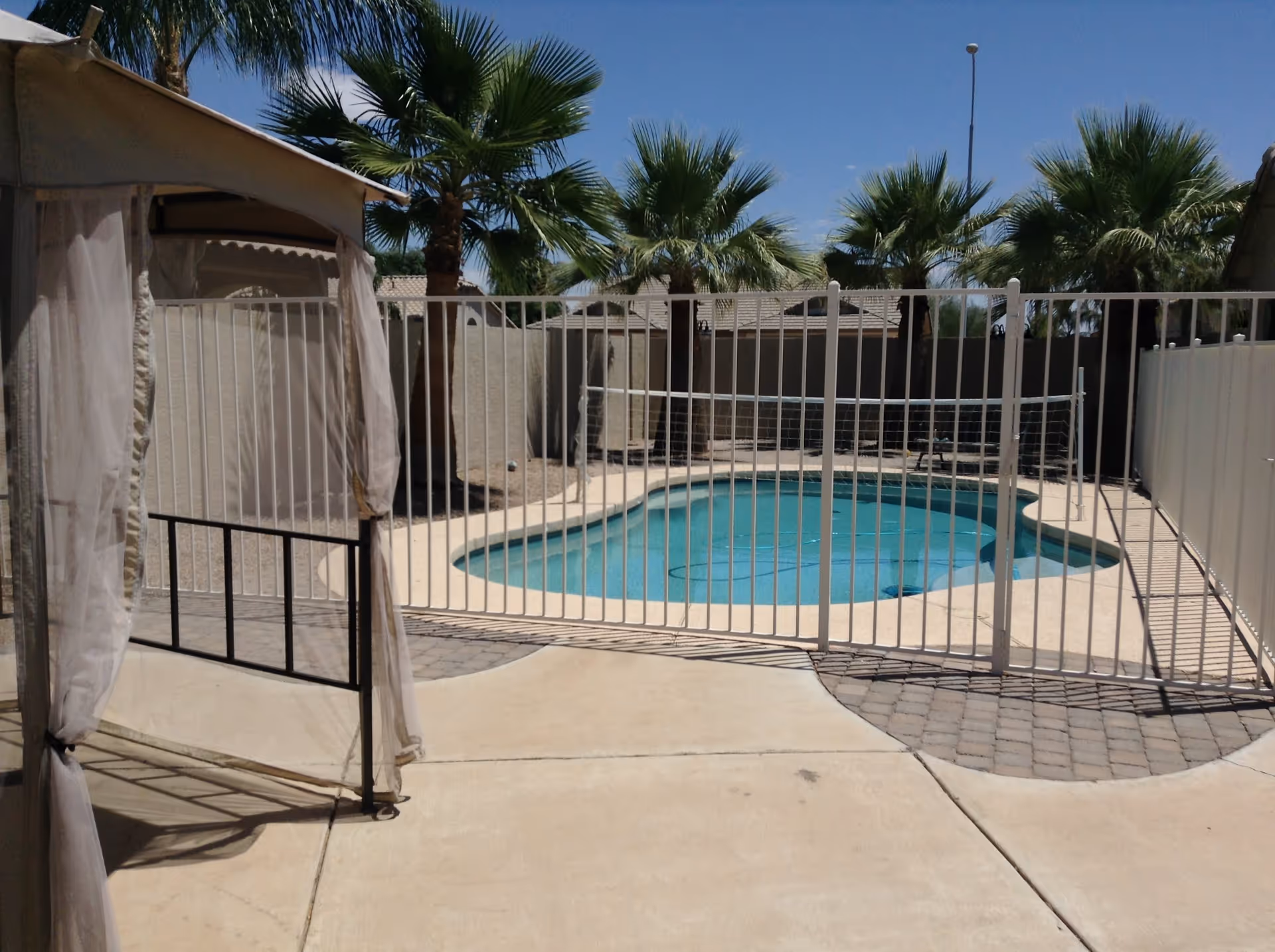 Outdoor swimming pool area enclosed by a white safety fence, with palm trees and a clear blue sky in the background. There is a beige canopy with sheer curtains on the left side and a paved walkway surrounding the pool.