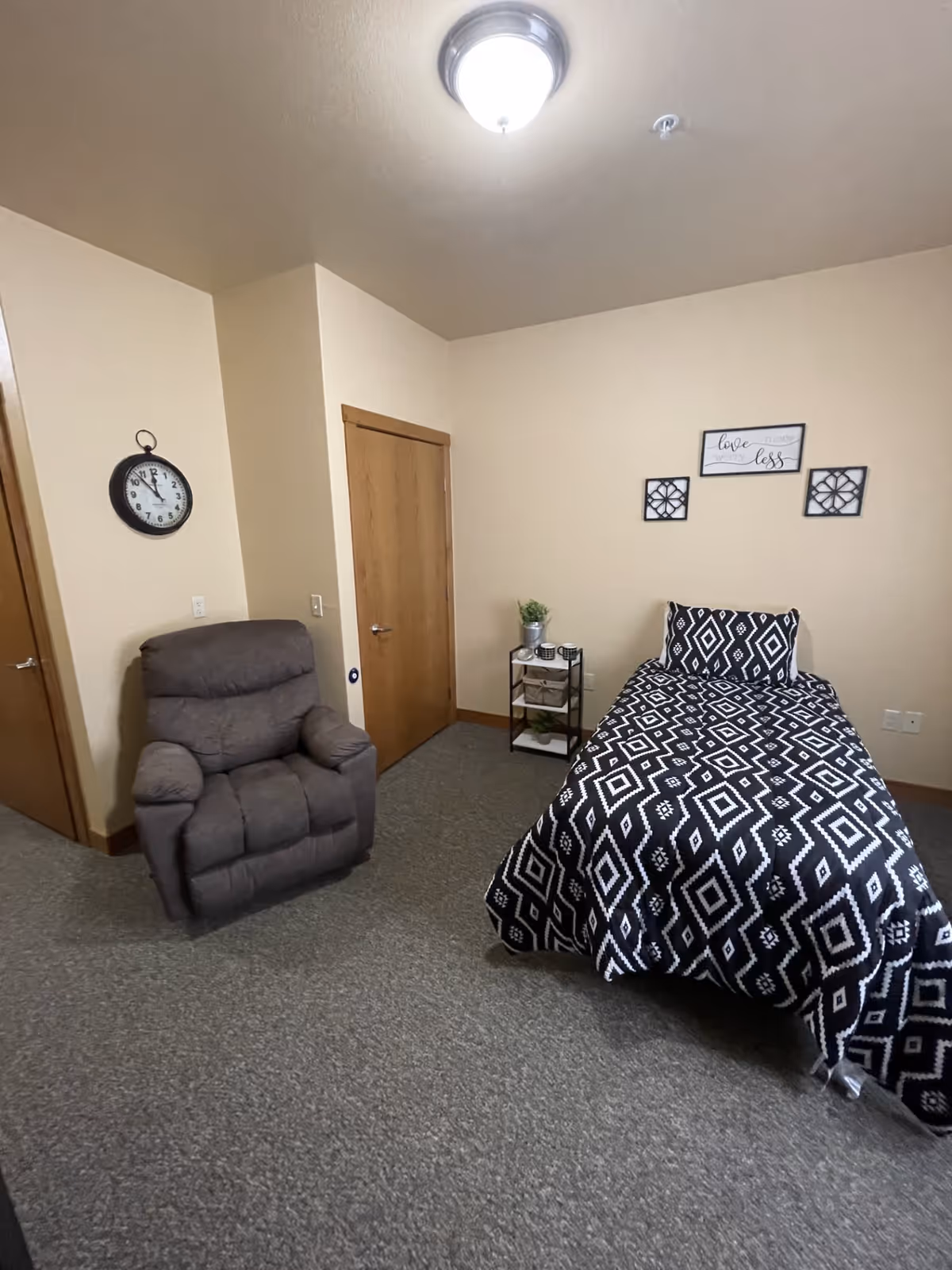 A small bedroom with a single bed covered in a black and white geometric patterned bedspread. Next to the bed is a small black metal side table with a plant and some decorative items. There is a gray recliner chair near two wooden doors. A round wall clock is mounted on the wall above the chair, and three framed wall decorations hang above the bed. The room has beige walls and gray carpeted flooring.