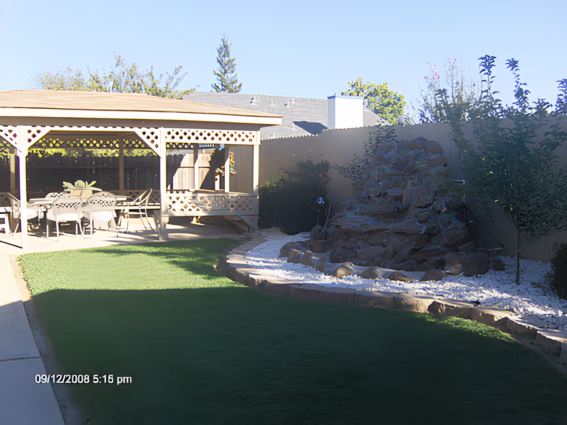 Outdoor garden area with a covered patio featuring a table and chairs, a green lawn, and a rock water feature surrounded by white gravel and plants along a wooden fence.