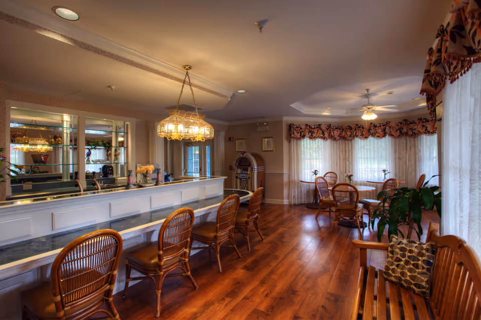 A cozy dining area in a senior living facility featuring a long counter with four wicker chairs, a chandelier above, and a mirrored wall behind the counter. In the background, there are round tables with wicker chairs, sheer curtains on large windows, a jukebox, and a ceiling fan. The room has warm wooden flooring and decorative valances on the windows.