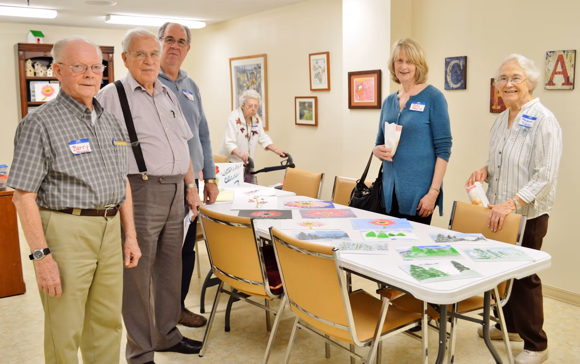 A group of older adults standing around a table in a communal room displaying drawings and paintings.
