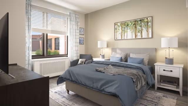 A well-lit bedroom with a large bed covered in blue bedding and gray pillows. There are two white nightstands on either side of the bed, each with a lamp. A framed picture of trees hangs above the bed. A window with blinds and patterned curtains lets in natural light. A dark chair with a pillow is placed near the window, and a dark dresser with a TV on top is visible in the foreground.