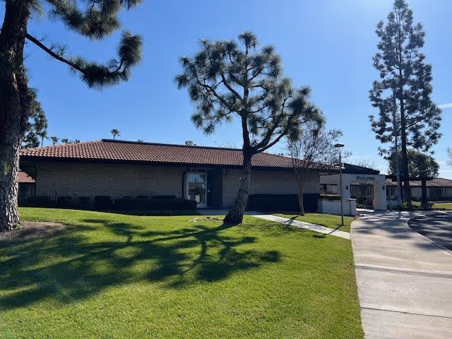 Exterior view of a single-story building with a tiled roof at a senior living community. The building is surrounded by green grass and several tall trees casting shadows on the lawn. A paved walkway leads to the entrance, where a sign reads 'Welcome'. The sky is clear and blue.