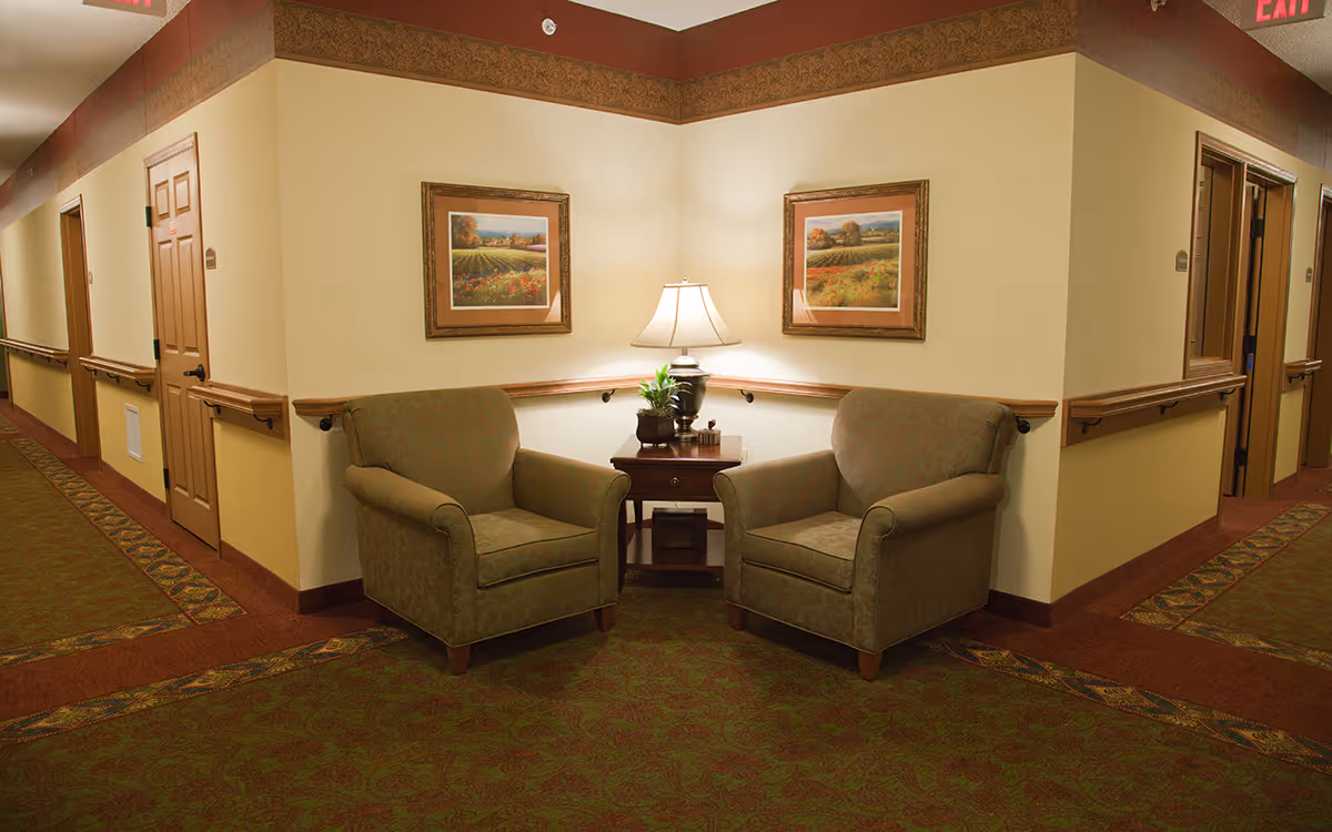Hallway seating area with two upholstered armchairs flanking a side table and lamp beneath framed paintings in a senior living facility corridor.