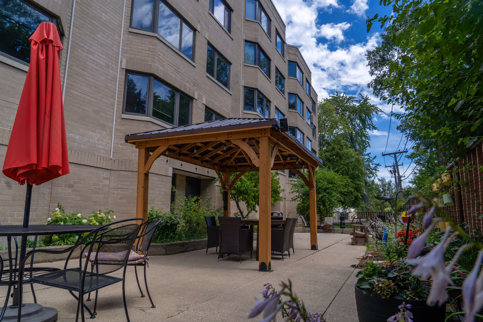 Outdoor patio area at Generations at Oakton Arms featuring a wooden gazebo with seating underneath, metal chairs and tables, a closed red umbrella, and surrounding greenery and flowers along a concrete pathway next to a multi-story brick building.