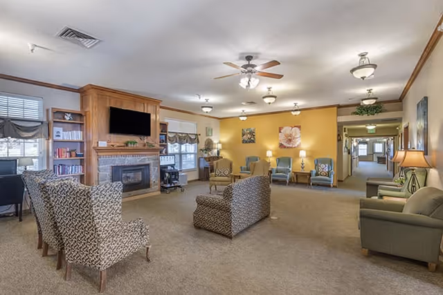 Spacious communal living room with upholstered chairs arranged around a fireplace and wall-mounted TV, warm lighting, and a yellow accent wall.