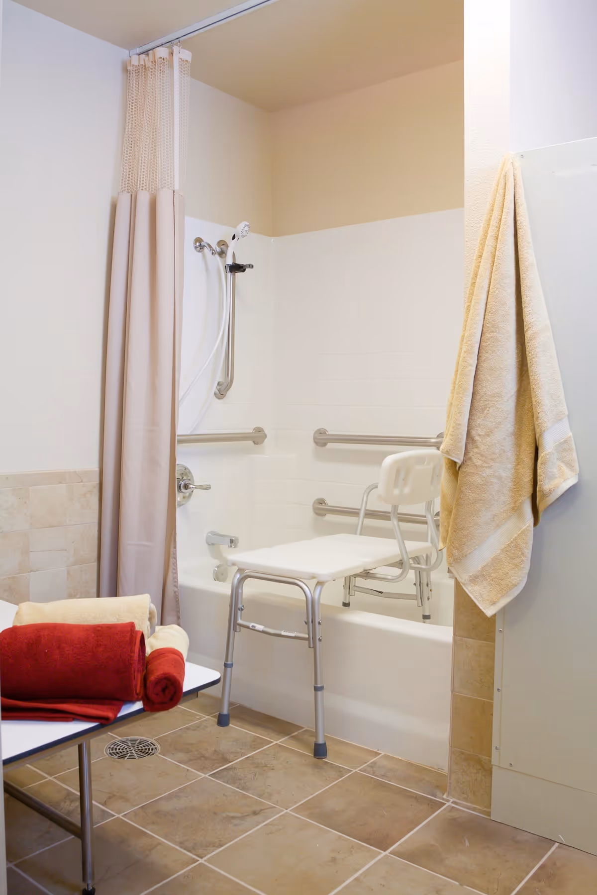 Bathroom with a bathtub fitted with a shower chair, grab bars, shower curtain, and towels.