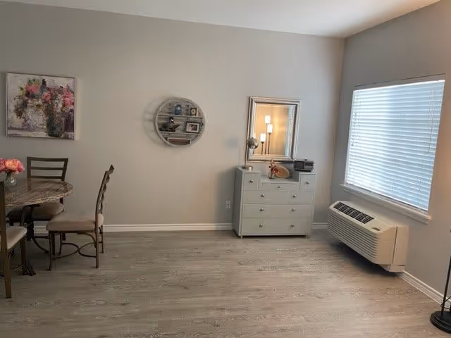 A room with light gray walls and wood flooring featuring a small dining table with chairs on the left, a round wall shelf with decorative items, a white dresser with a mirror and lamp on top, and a window with blinds above a wall-mounted air conditioning unit on the right.
