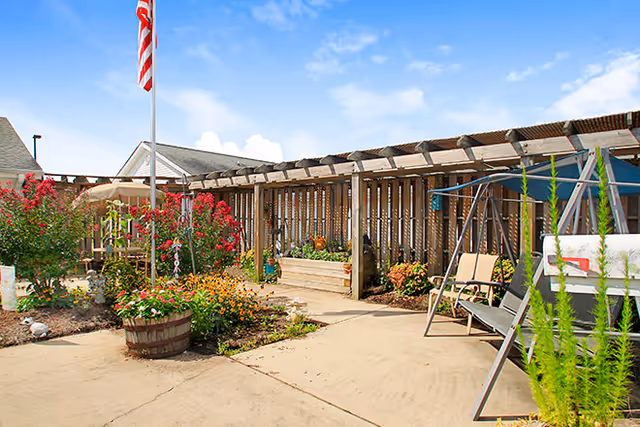 Sunny courtyard with a wooden pergola, flower beds, an American flag, and outdoor seating and swings.