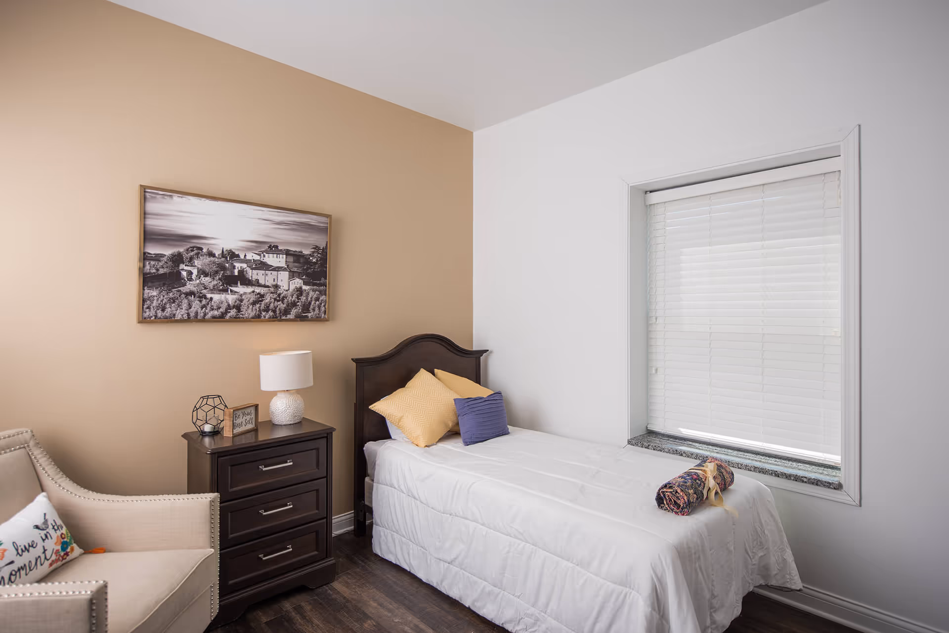 A small bedroom with a single bed covered in white bedding and two pillows, one yellow and one purple. Next to the bed is a dark wooden nightstand with a white lamp, a decorative object, and a small framed sign. There is a beige armchair with a colorful pillow that says 'live in the moment'. A large window with white blinds is on the right wall, and a framed black and white landscape photograph hangs on the beige wall behind the bed.