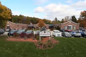 Front exterior view of Oakdale Rehabilitation & Skilled Nursing Center building with a parking lot filled with cars in front, surrounded by trees with autumn foliage under a partly cloudy sky.