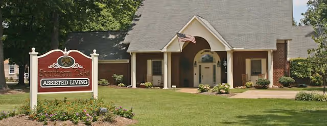 Exterior view of Southern Oaks Assisted Living facility showing a large building with a steep roof, an American flag near the entrance, and a landscaped lawn with bushes and flowers. A red and white sign in the foreground reads 'Southern Oaks Assisted Living.'